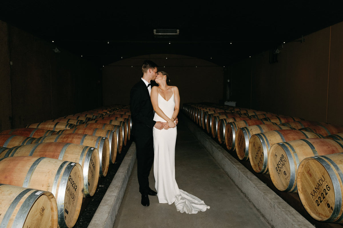 A bride and a groom in a black suit kiss in a dimly lit wine cellar aisle lined with wooden barrels.