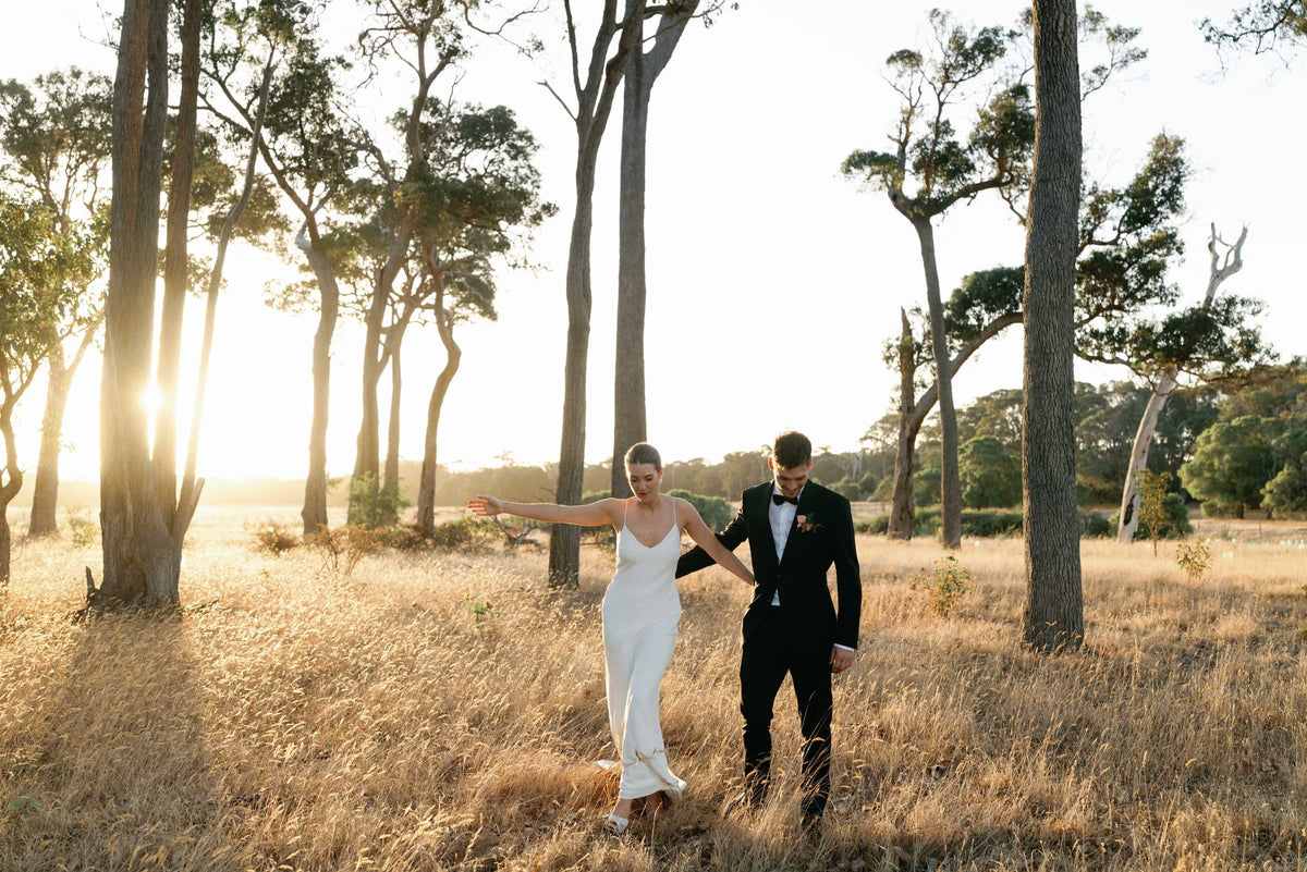 A bride and a groom in a black suit walk arm in arm through a sunlit grassy field with tall trees.