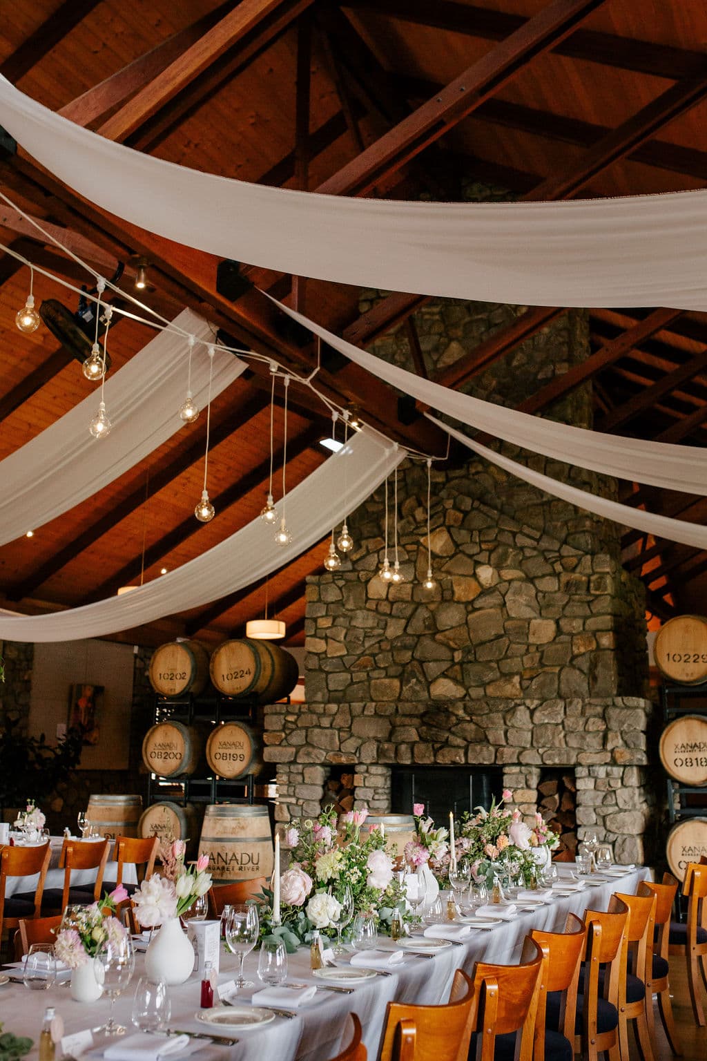 Xanadu event space with a stone fireplace, exposed wooden ceiling beams, draped white fabric, hanging string lights, and a long dining table set with white linens, floral centerpieces, and wooden chairs.