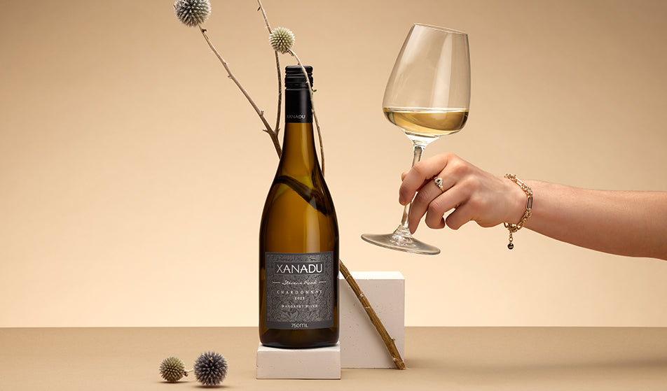 Bottle of Xanadu Boodjidup Brook Chardonnay beside a glass of white wine held by hand, with dried flowers