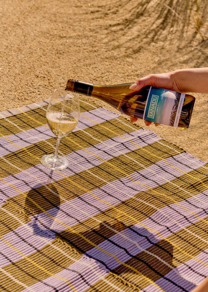 Hand pouring a bottle of Xanadu sparkling wine into a glass on a plaid picnic blanket on the sand