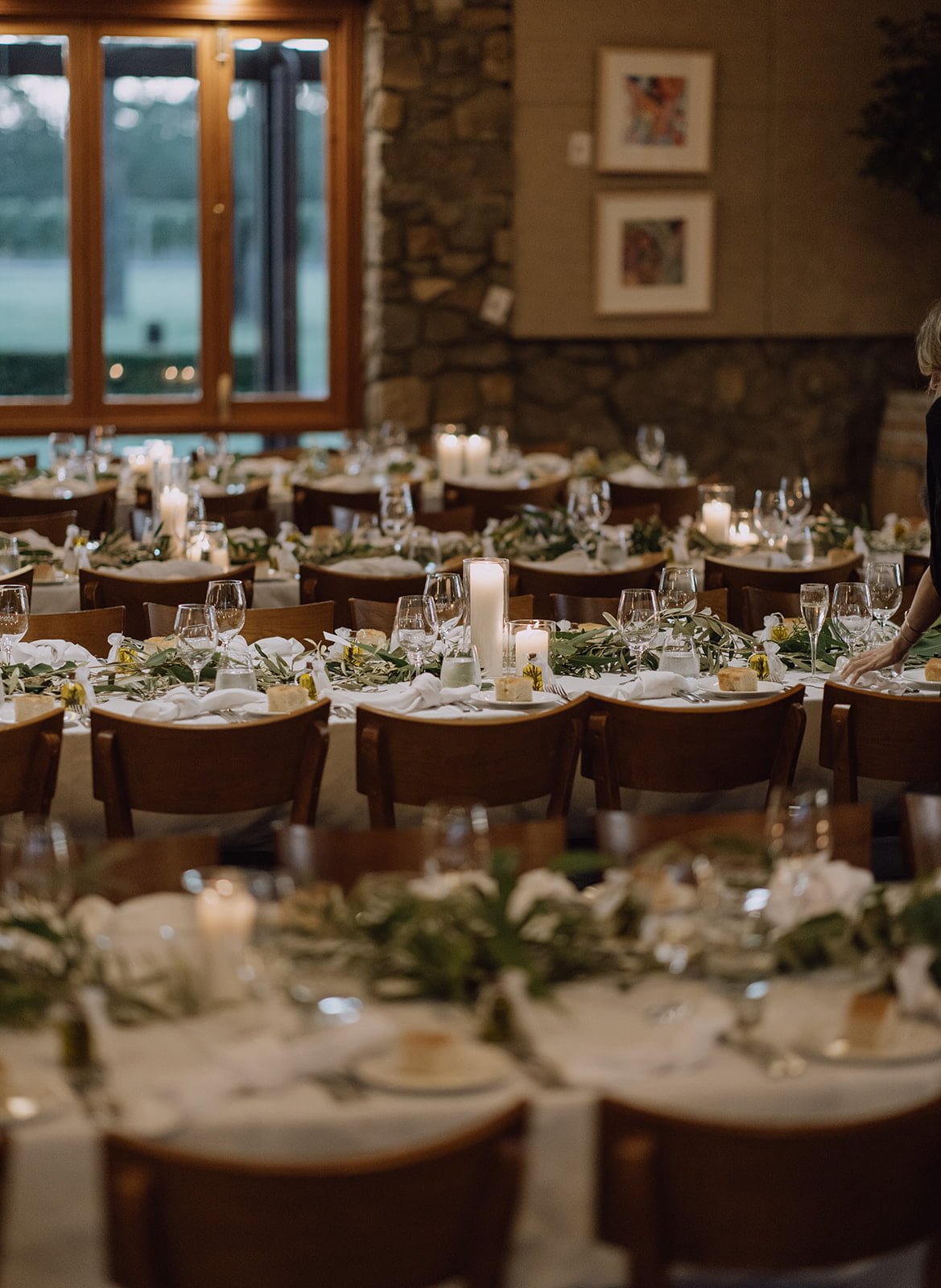 Xanadu wedding reception space with multiple round tables set for dining, featuring white tablecloths, green floral centerpieces, lit candles, and wooden chairs. A stone wall and large windows are visible in the background.