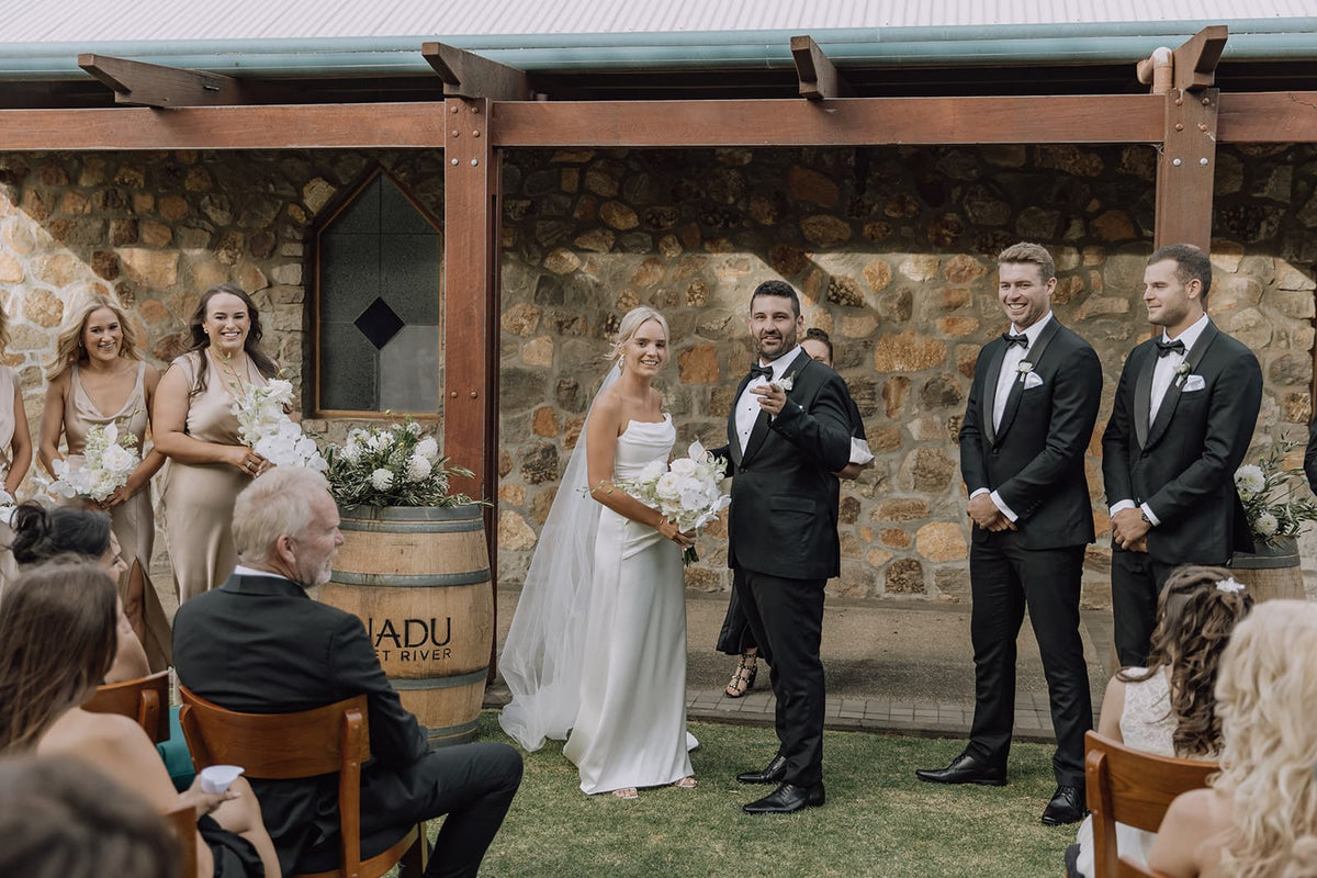 Xanadu wedding ceremony scene outdoors under a wooden pergola, with a bride and groom standing on grass, surrounded by guests and bridesmaids. 