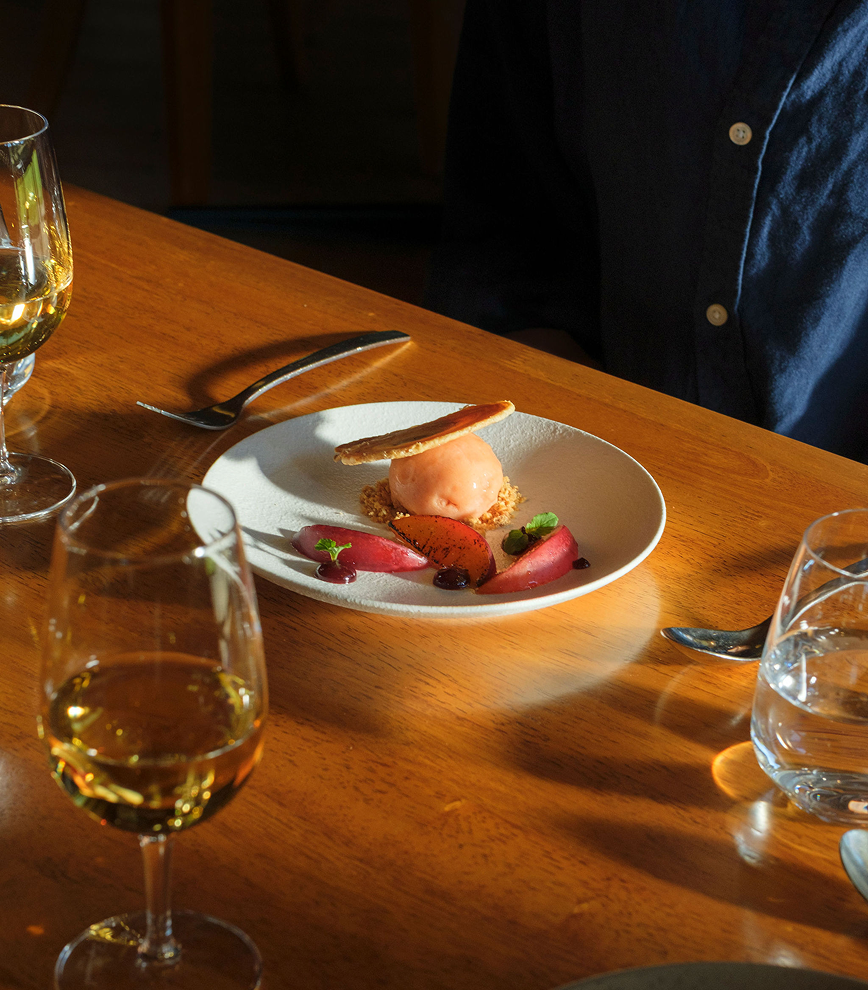 Restaurant dessert of ice cream and stone fruit on a plate beside glasses of white wine on a wooden table