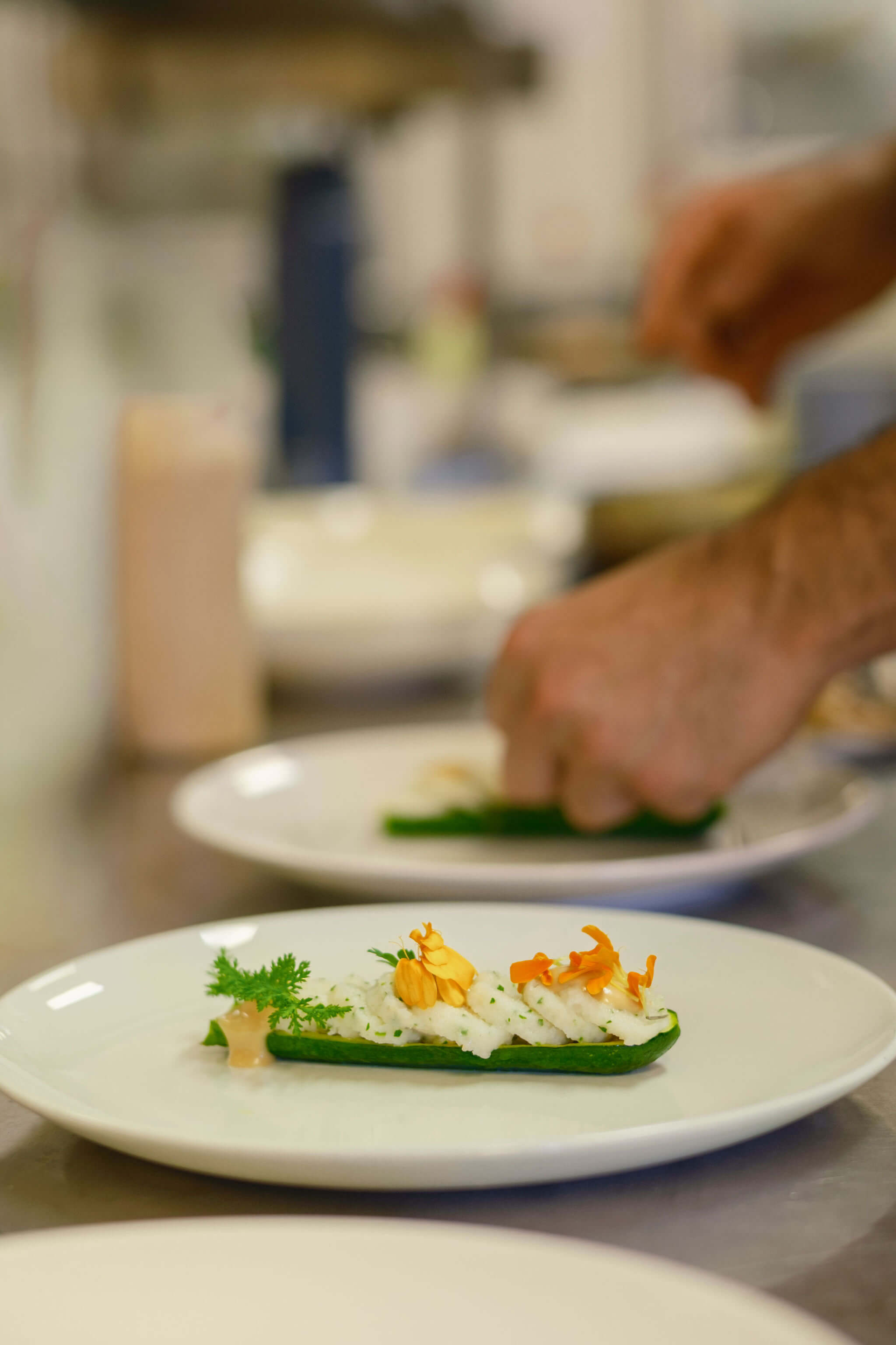 Chef plating a dish of fish over cucumber ribbons with edible flowers in the restaurant kitchen