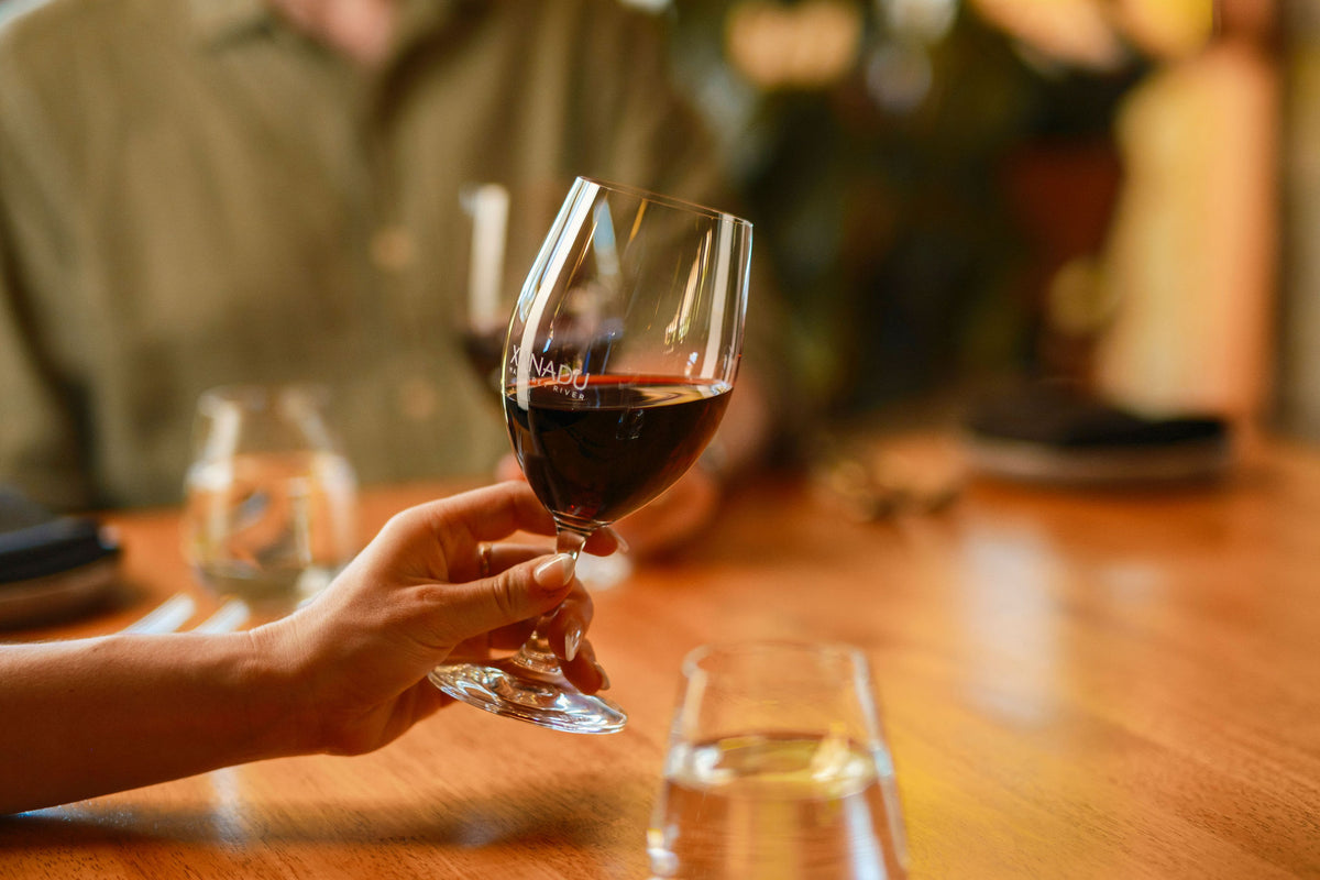 Hand holding a glass of red wine with the Xanadu logo, at a restaurant table