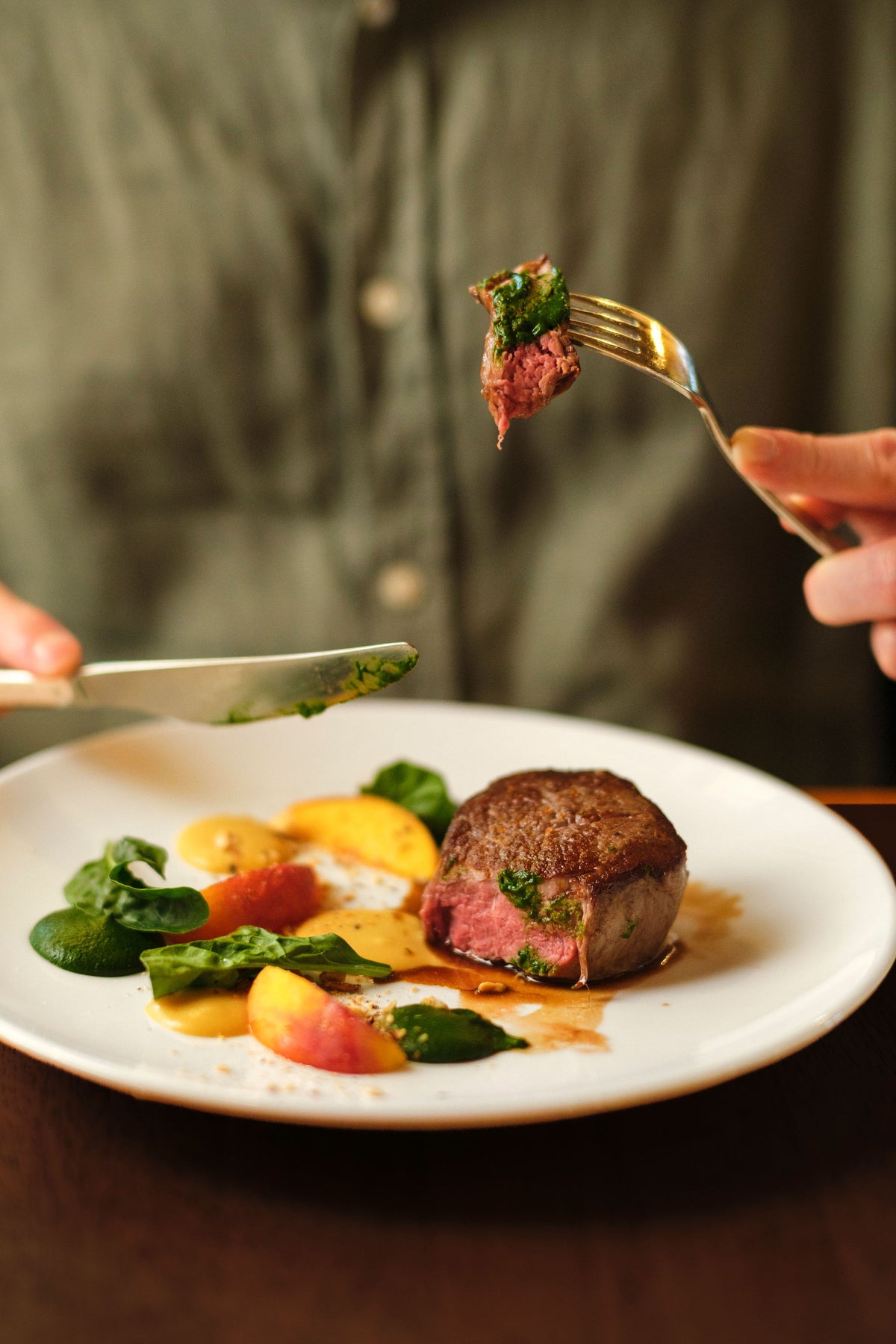 Diner cutting into a seared steak served with peaches and greens on a white plate