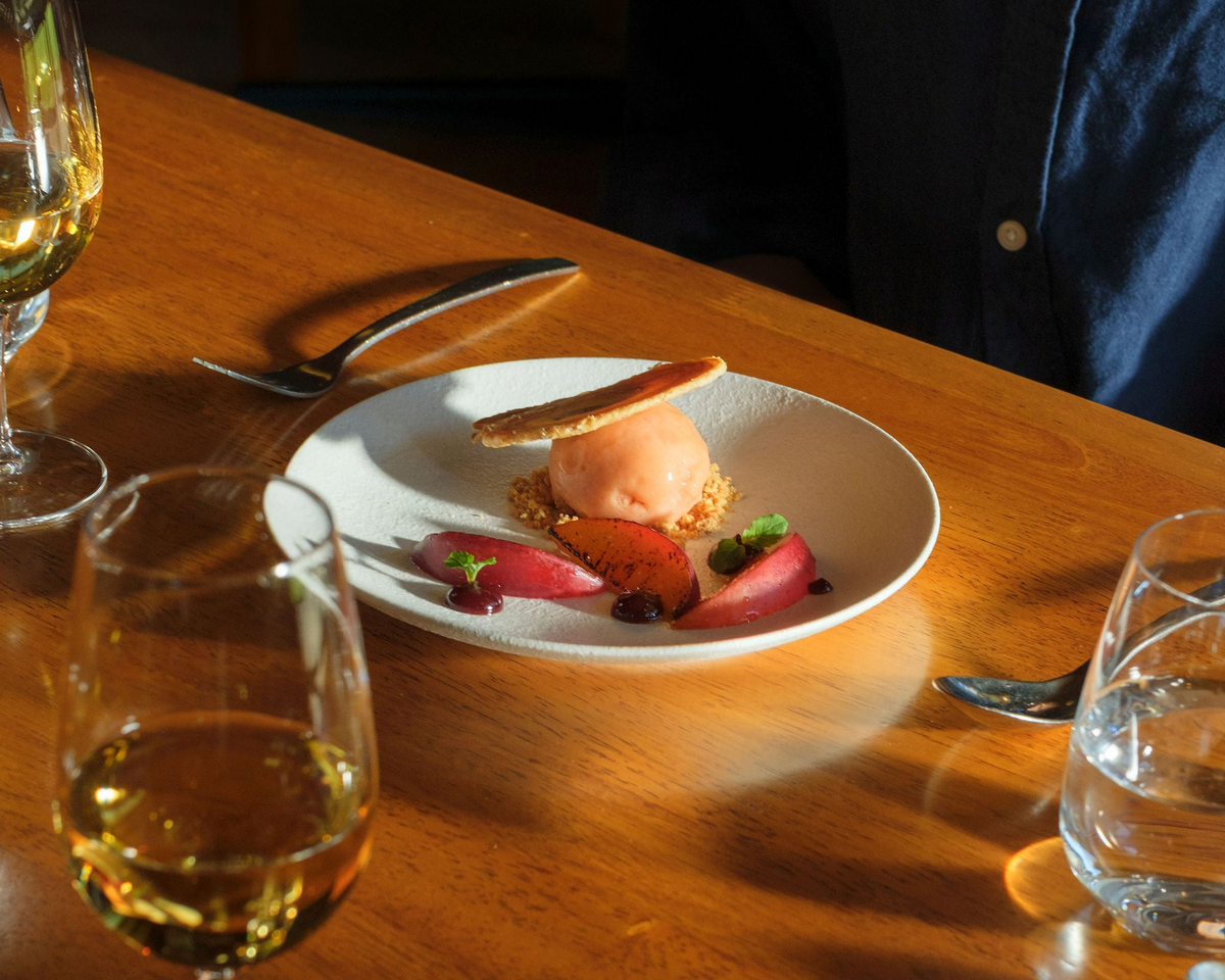 Restaurant dessert of ice cream and stone fruit on a plate beside glasses of white wine on a wooden table