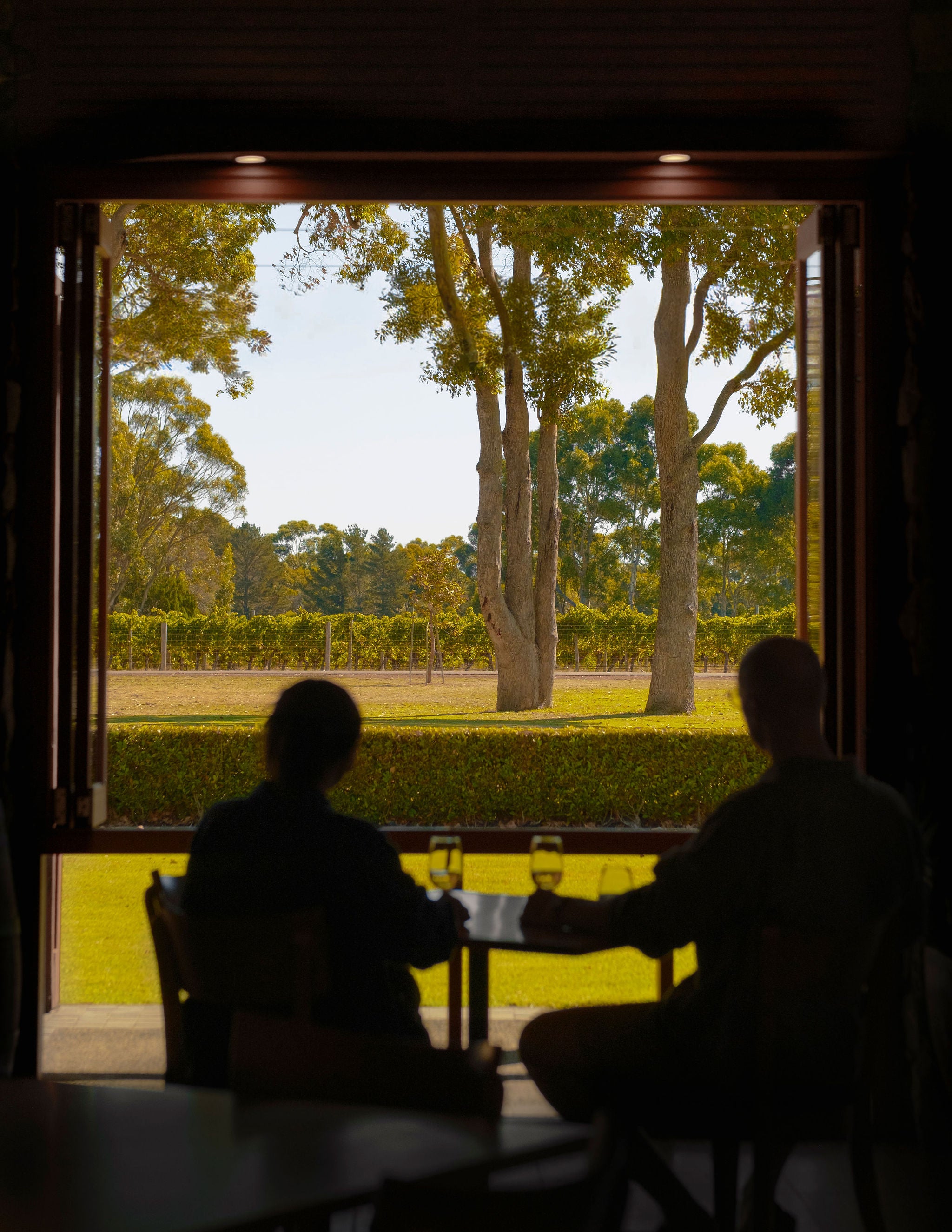 Two diners silhouetted at a table with wine glasses, looking out over the vineyard through an open window