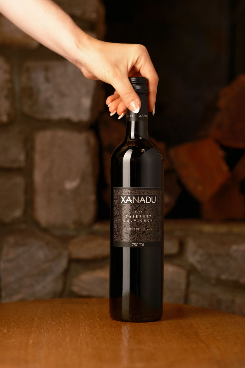 Hand reaching for a bottle of Xanadu Cabernet Sauvignon on a table beside a stone fireplace
