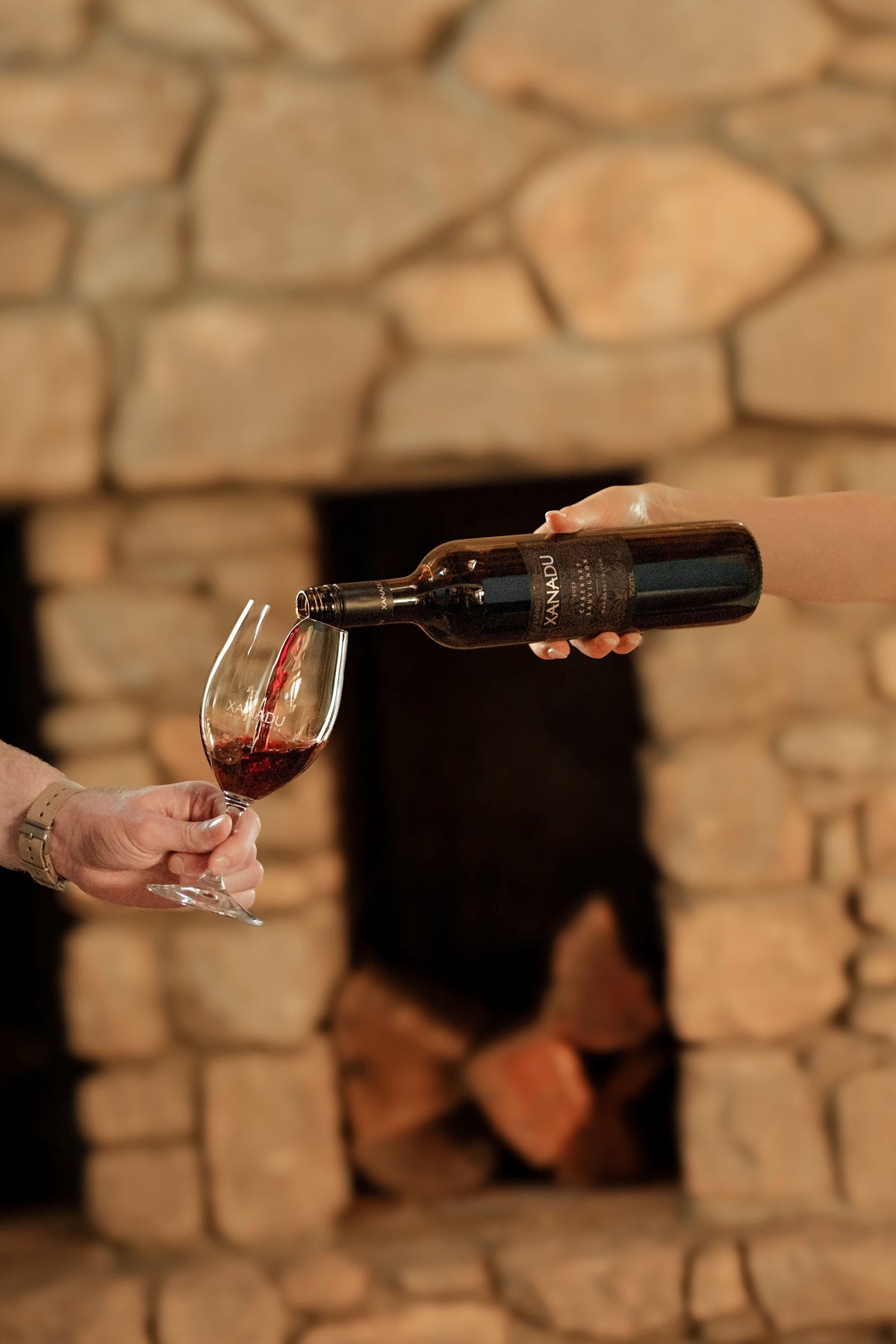 Hand pouring a bottle of Xanadu wine into a glass of red wine in front of a stone fireplace