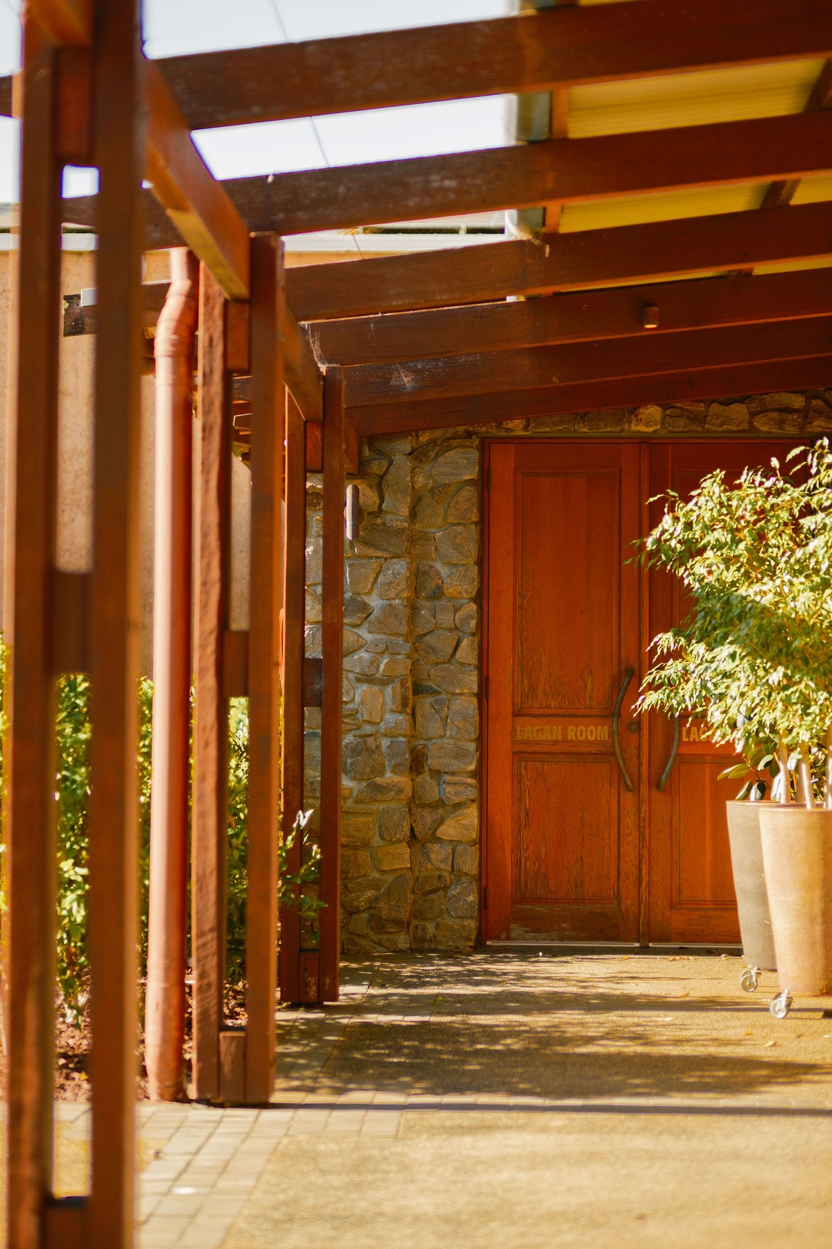 Timber-covered walkway leading to a stone entrance with wooden doors and potted plants