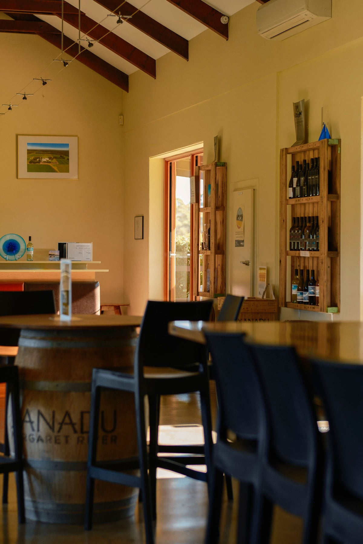 Interior of the Xanadu cellar door with a wine barrel table, bar stools, and shelves stocked with wine bottles