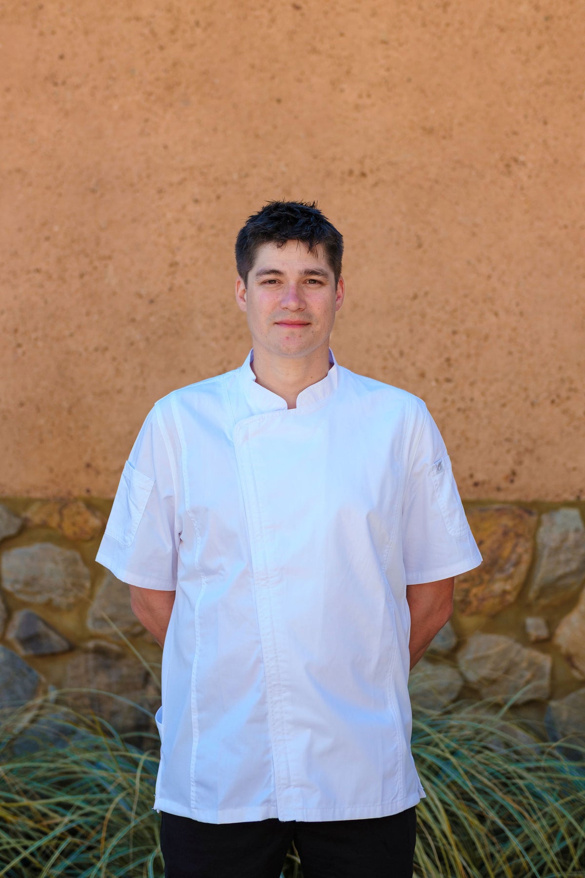 Portrait of a smiling Xanadu chef in white chef's jacket standing against a stone and rendered wall
