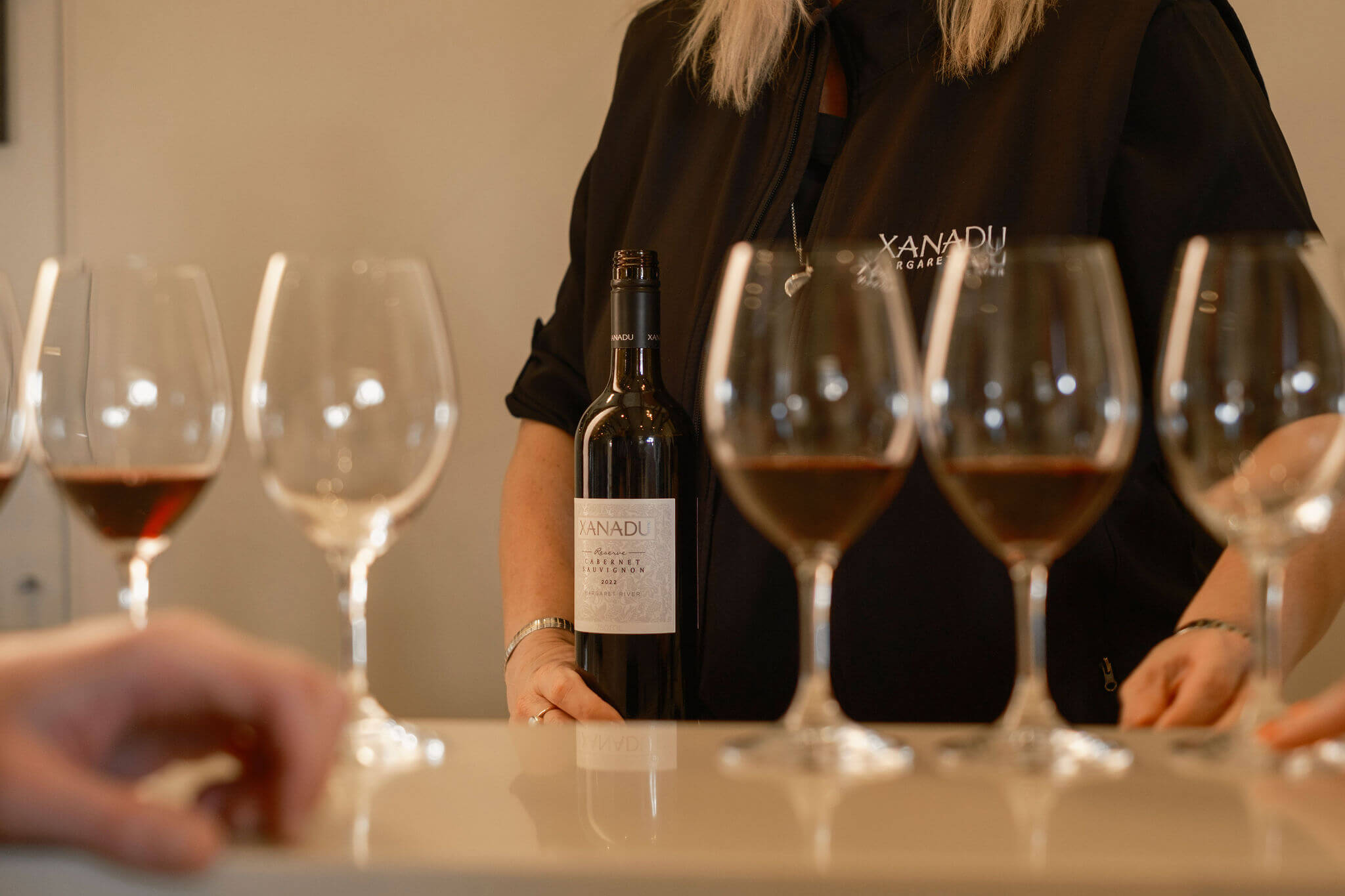 Xanadu team member in branded vest holding a bottle of Reserve Cabernet Sauvignon behind a row of wine tasting glasses