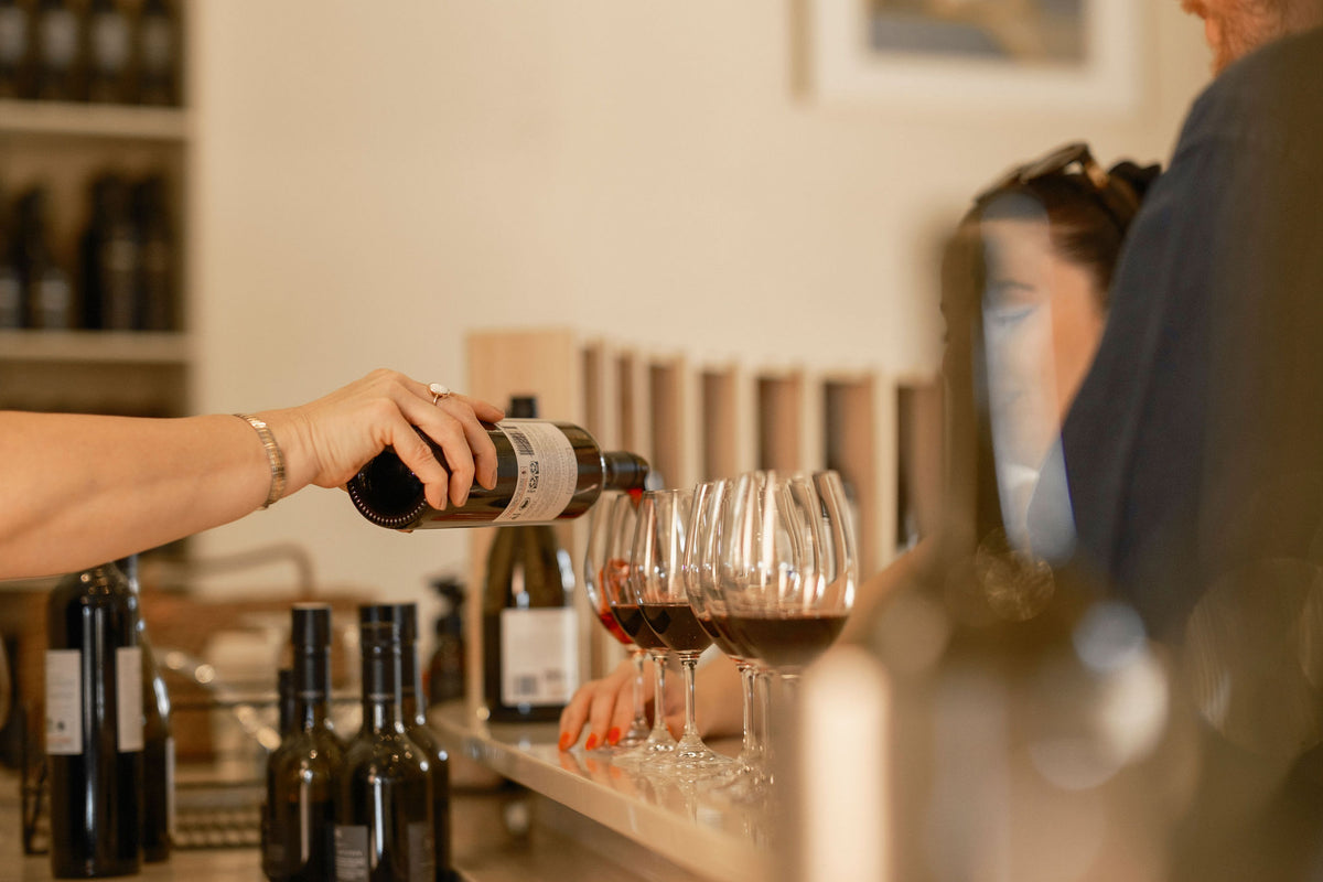 Hand pouring a bottle of Xanadu wine into a row of glasses at the cellar door tasting bar
