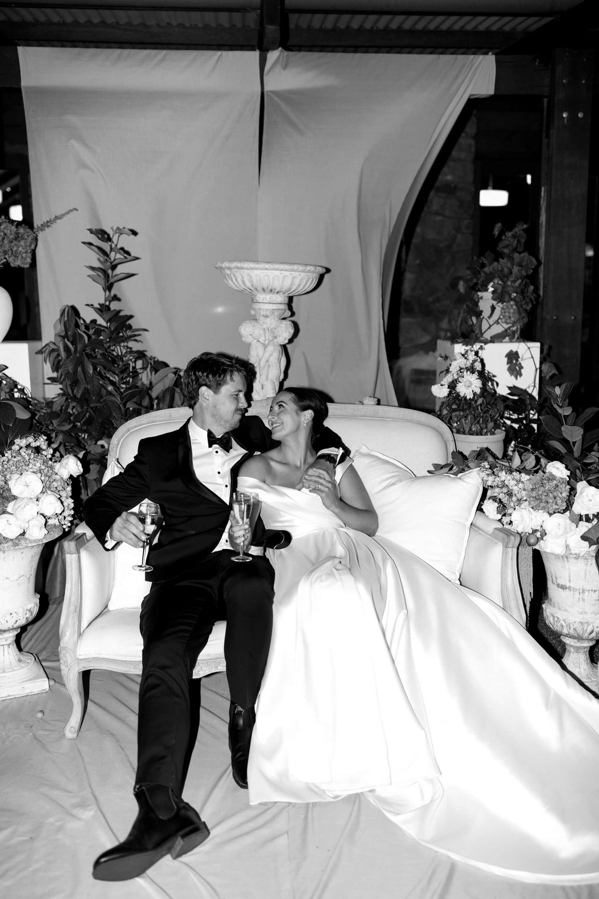 Black and white wedding photograph of a bride and groom sitting together on a white armchair, with flowers, draped fabric, and event decor in the background.