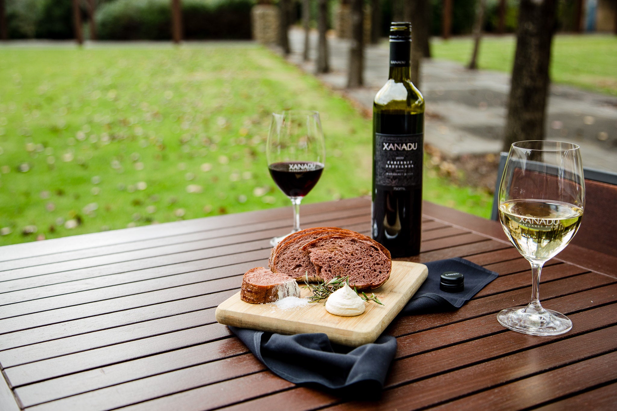 Bottle of Xanadu Cabernet Sauvignon and glasses of red and white wine beside a board of bread and whipped butter on an outdoor table