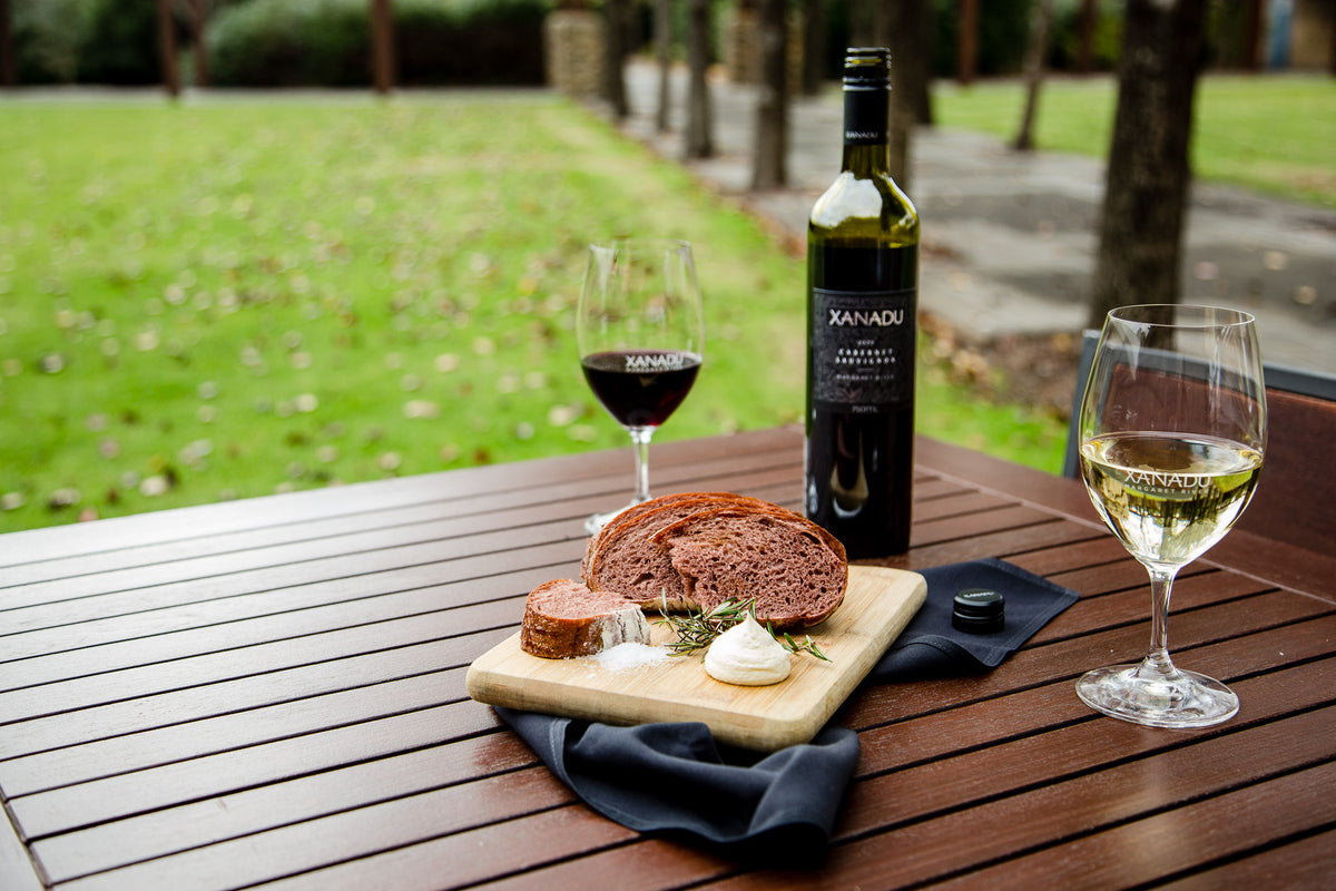 Bottle of Xanadu Cabernet Sauvignon and glasses of red and white wine beside a board of bread and whipped butter on an outdoor table
