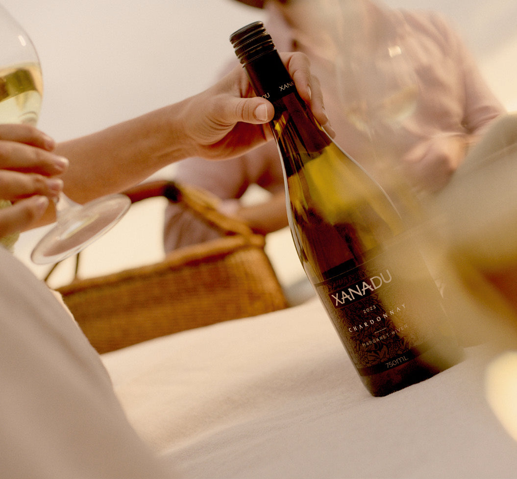 Hand placing a bottle of Xanadu Chardonnay near a wicker picnic basket, with a person holding a glass of white wine