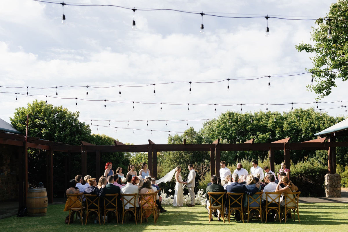 Outdoor wedding reception scene under a pergola with guests seated on a grassy lawn, string lights overhead, trees in the background, and a bride in a white gown standing near the altar.