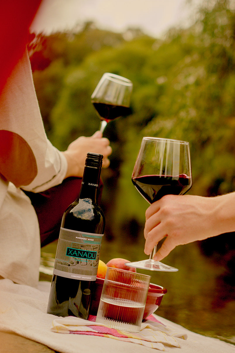 Bottle of Xanadu Cabernet Sauvignon beside a glass of red wine, peaches, and a picnic blanket by the water