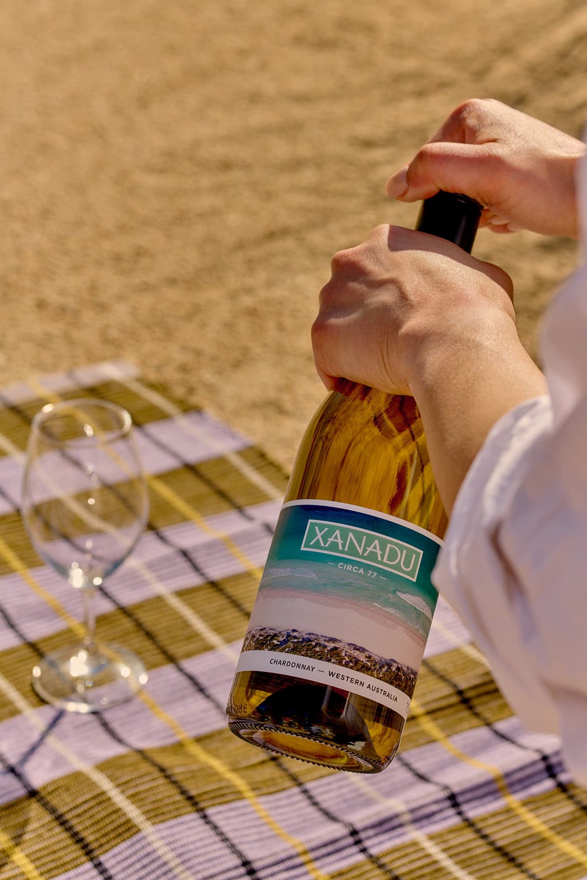 Hands opening a bottle of Xanadu Circa 77 Chardonnay on a picnic blanket on the sand