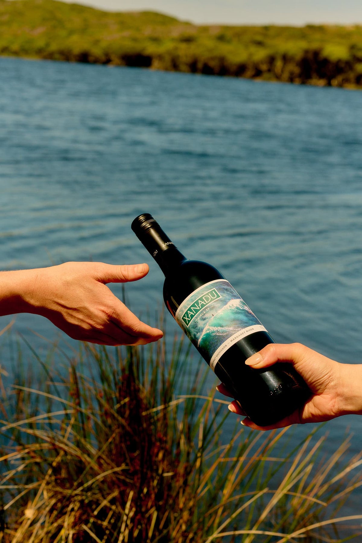 Two hands passing a bottle of Xanadu Cabernet Sauvignon beside a river with dune grass