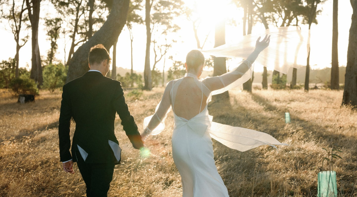 Bride and groom walking hand in hand through sunlit trees, the bride's veil billowing in the breeze