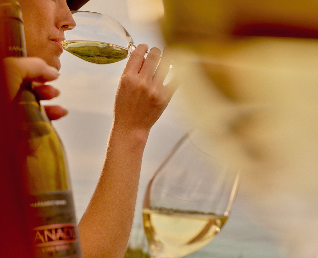Person tasting a glass of white wine beside a bottle of Xanadu wine, ocean in the background