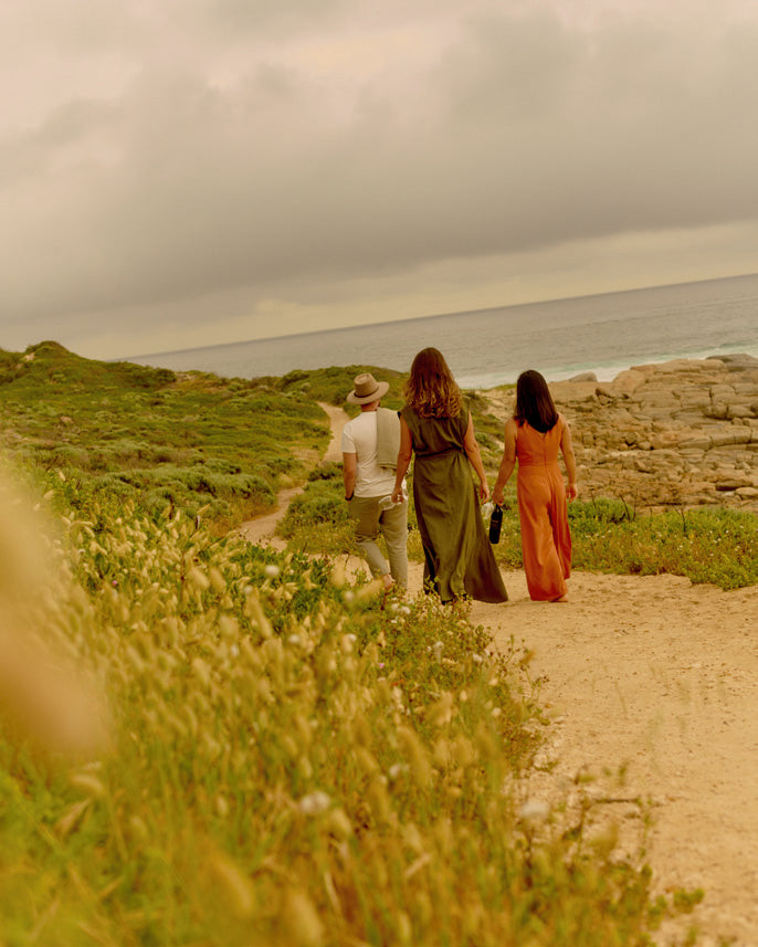 Three people walking away along a coastal path toward the ocean, one carrying a bottle of wine