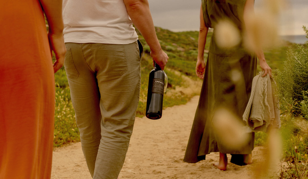 Three people walking along a coastal path, one carrying a bottle of Xanadu wine