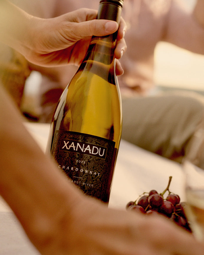 Hand holding a bottle of Xanadu Chardonnay at a table with grapes and a glass of white wine, blurred figure in the background