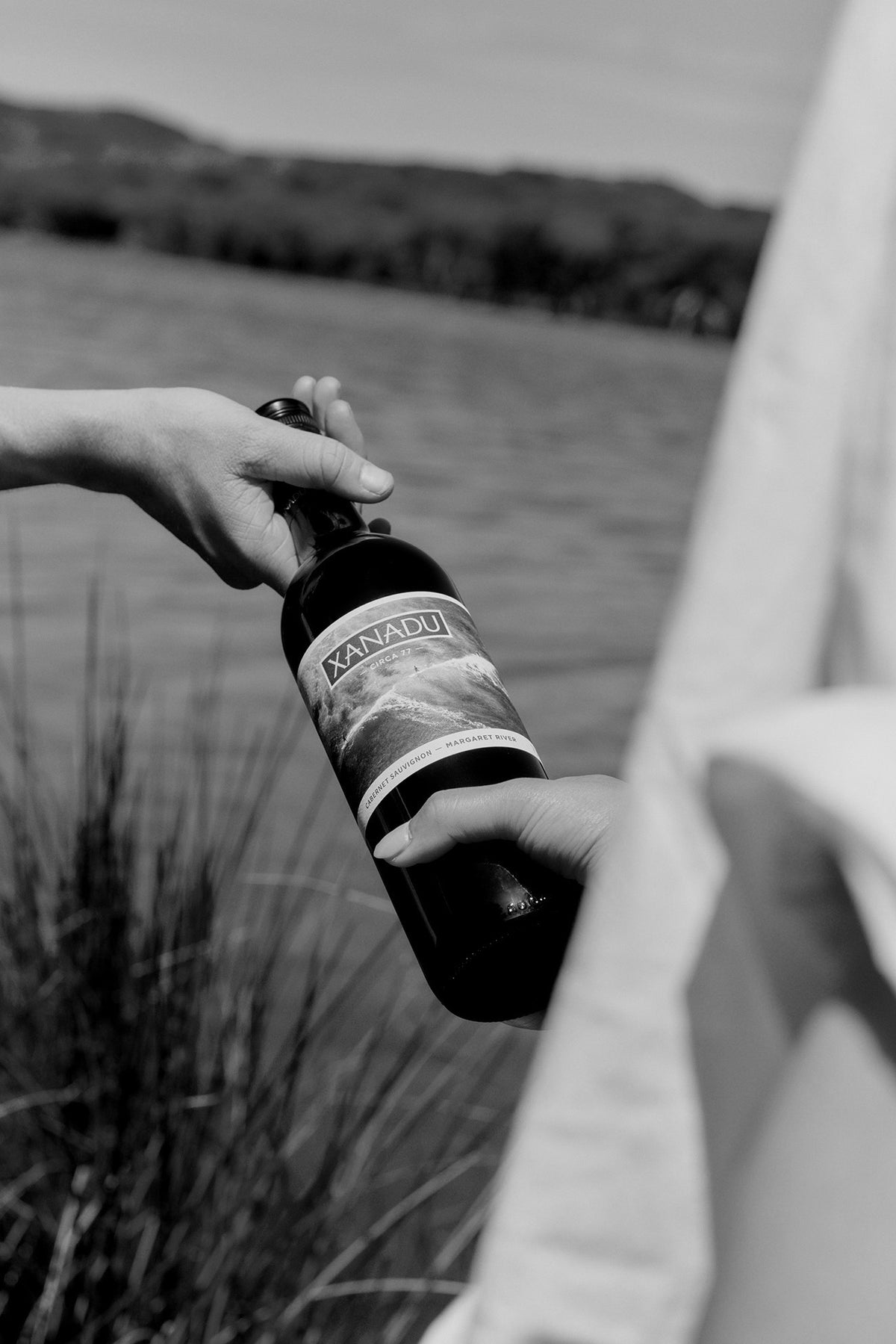 Black and white photo of a hand holding a bottle of Xanadu wine beside a lake