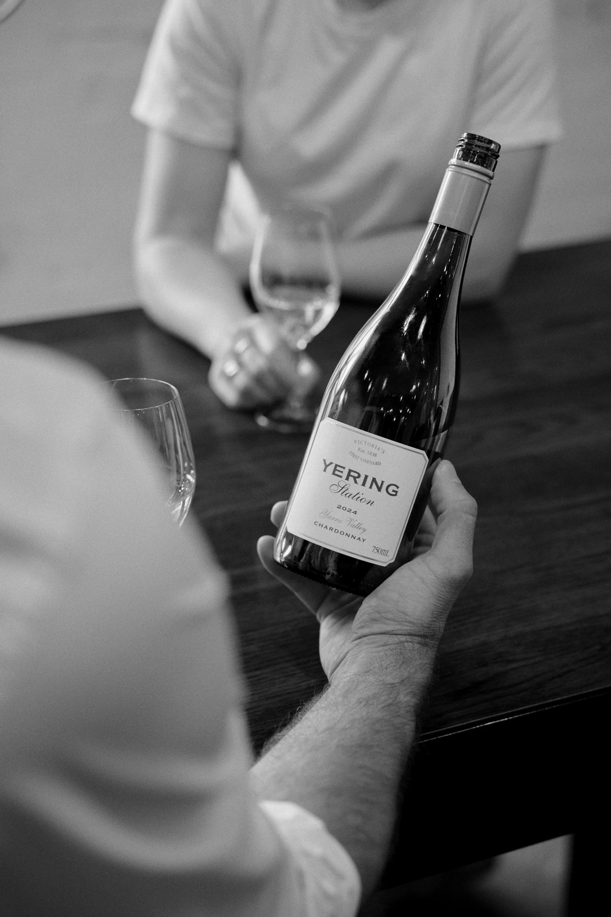 Black and white photo of a hand holding a bottle of Yering Station Chardonnay across a table from a seated guest